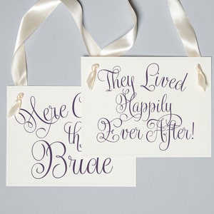 May include: Two ivory wedding signs with cream satin ribbon handles. One sign reads "Here Comes the Bride" in purple script, the other says "They Lived Happily Ever After!"
