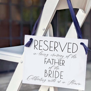 Può includere: Sedia bianca con un cartello che recita "RESERVED in loving memory of the FATHER of the BRIDE Celebrating with us in Heaven". Il cartello è attaccato alla sedia con un nastro blu.