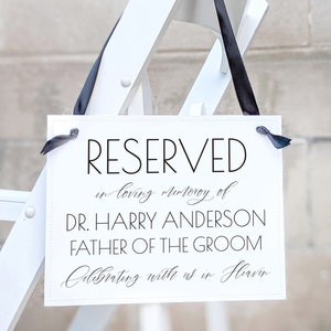 Op de afbeelding: Een wit bord met zwarte tekst dat "RESERVED in loving memory of DR. HARRY ANDERSON FATHER OF THE GROOM Celebrating with us in Heaven." zegt.