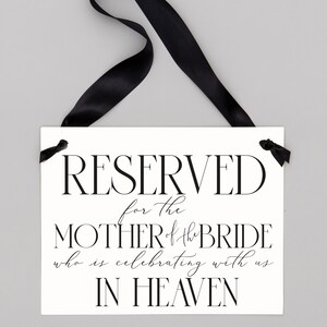 May include: White sign with black text that reads "RESERVED for the MOTHER of the BRIDE who is celebrating with us IN HEAVEN". The sign is hanging from a black ribbon.
