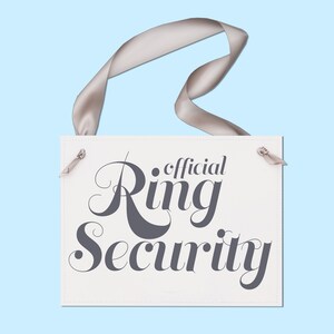 May include: A rectangular sign with the words "official Ring Security" in dark gray script. The sign is white with a stitched border and a gray ribbon for hanging. The background is a light blue.
