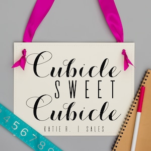 May include: White sign with black cursive text that reads "Cubicle Sweet Cubicle" and "Katie R. | Sales". The sign is hanging from pink ribbon.