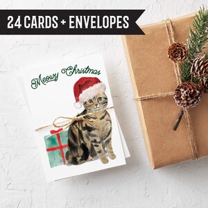 May include: Christmas greeting cards featuring a watercolor illustration of a tabby cat wearing a Santa hat. The cards are tied with twine and include the text "Meowy Christmas." The image also shows a wrapped gift and the text "24 CARDS + ENVELOPES."