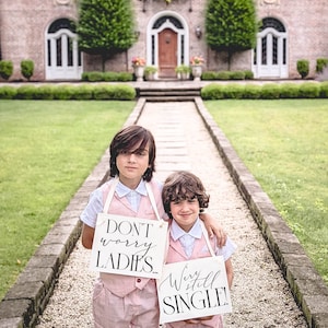 May include: Two young people in matching pink suits stand on a stone path, holding signs. One sign reads "DON'T worry LADIES..." and the other says "We're still SINGLE!". The background features a large brick house and green lawn.