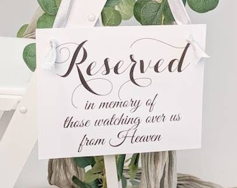 May include: A white rectangular sign with black script text that reads Reserved in memory of those watching over us from Heaven. The sign is suspended by white ribbons from a white wooden frame adorned with green eucalyptus leaves and grey fabric.