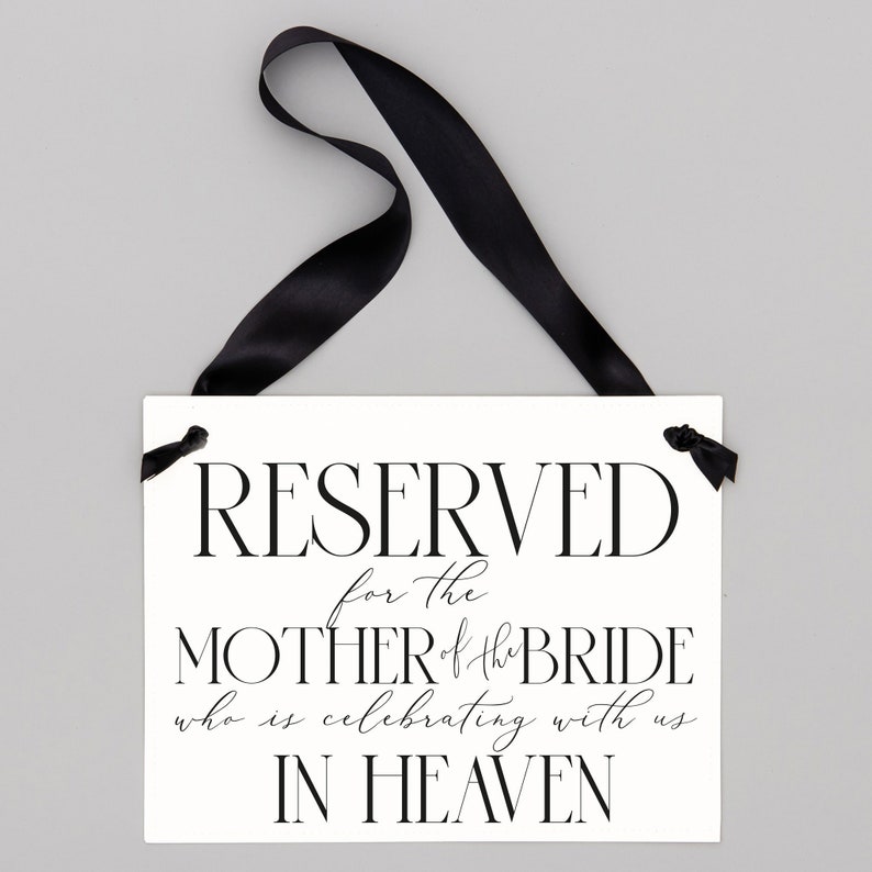 May include: White sign with black text that reads "RESERVED for the MOTHER of the BRIDE who is celebrating with us IN HEAVEN". The sign is hanging from a black ribbon.