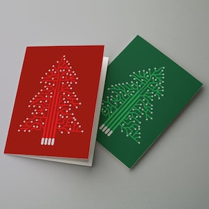Puede incluir: Dos tarjetas de felicitación plegadas con un diseño de árbol de Navidad. Una tarjeta es roja con un árbol de placa de circuito blanco, y la otra es verde con un árbol de placa de circuito blanco. Las tarjetas están sobre una superficie gris claro.