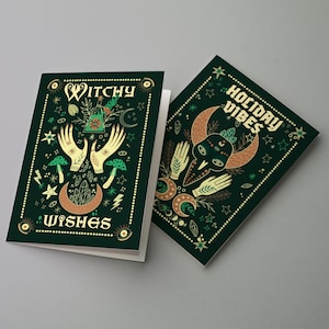 May include: Two dark green greeting cards with gold accents. One card reads "Witchy Wishes" with illustrations of hands, mushrooms, and moons. The other card says "Holiday Vibes" with similar imagery. Both cards have a border of small dots.