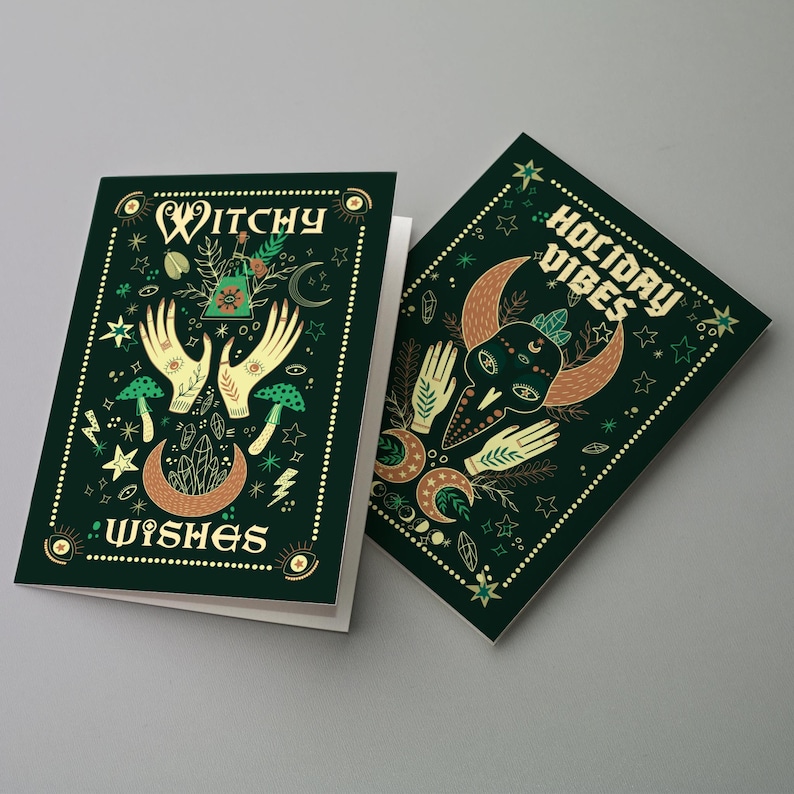 May include: Two dark green greeting cards with gold accents. One card reads "Witchy Wishes" with illustrations of hands, mushrooms, and moons. The other card says "Holiday Vibes" with similar imagery. Both cards have a border of small dots.