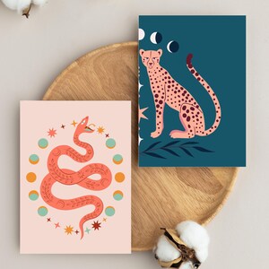 Può includere: Due stampe artistiche rettangolari. Una raffigura un serpente color corallo circondato da un cerchio di stelle e fasi lunari su sfondo rosa chiaro. La seconda mostra un ghepardo rosa su sfondo verde acqua scuro con fasi lunari e stelle.