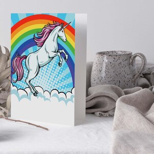 Può includere: Un biglietto d'auguri con un unicorno bianco dalla criniera e coda rosa, che si impenna davanti a un vivace sfondo arcobaleno. Il design presenta un motivo a raggiera blu con punti bianchi e nuvole bianche stilizzate sul bordo inferiore.
