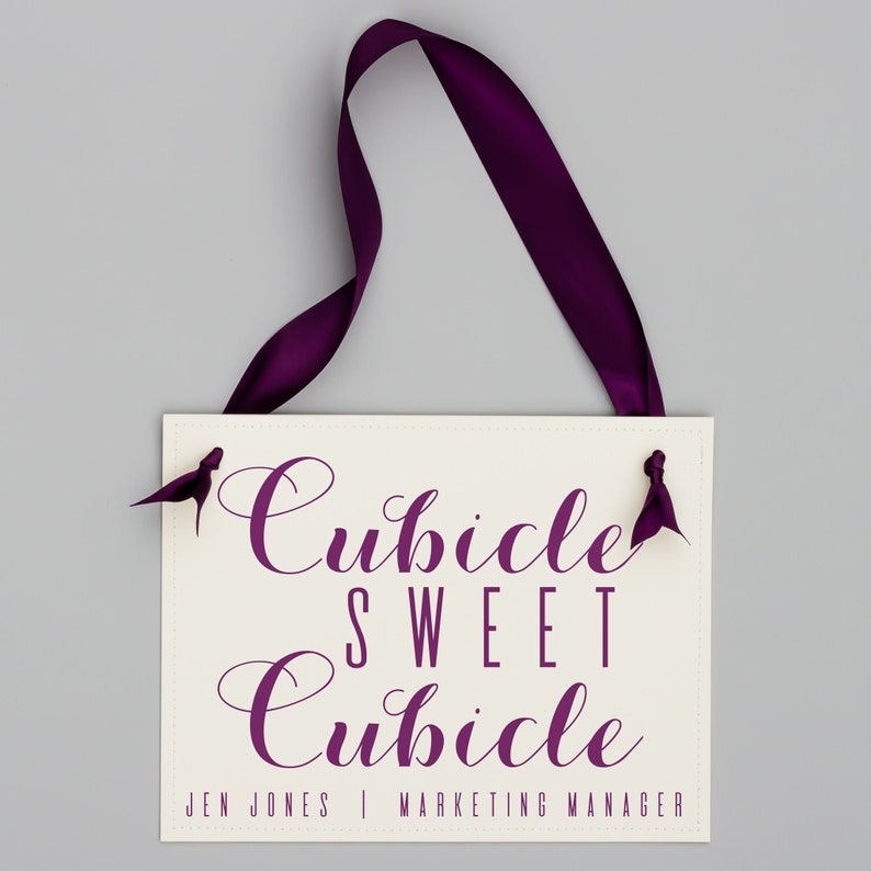 May include: A white sign with purple text that reads "Cubicle Sweet Cubicle" and "Jen Jones | Marketing Manager". The sign is hanging from a purple ribbon.