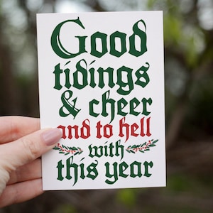 May include: A white greeting card with the text "Good tidings & cheer and to hell with this year" in green and red lettering. The card is held by a person's hand against a blurred green background.