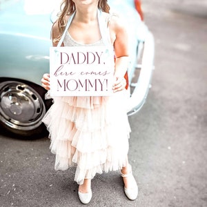 May include: A young girl in a white dress holds a sign that says "Daddy, here comes Mommy!" in burgundy lettering. The sign is white with a light blue ribbon.