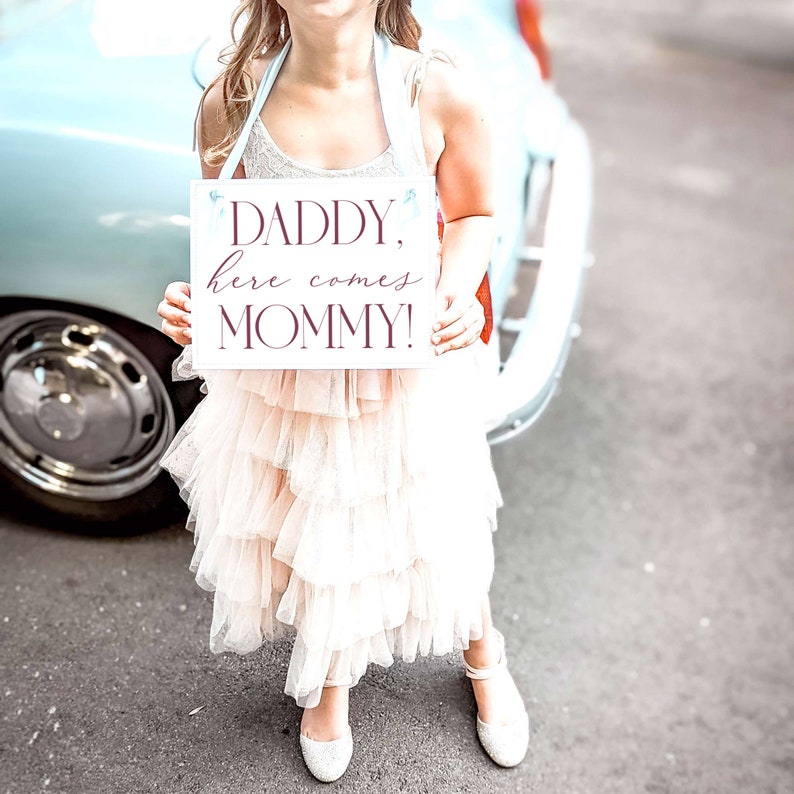 May include: A young girl in a white dress holds a sign that says "Daddy, here comes Mommy!" in burgundy lettering. The sign is white with a light blue ribbon.