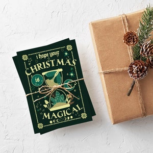 May include: Dark green Christmas cards tied with twine, featuring the text "I hope your Christmas is Magical" in a whimsical font. The cards have an hourglass and botanical illustrations. A wrapped gift with pinecones and greenery is in the background.