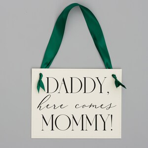 May include: White sign with black text that reads "Daddy, here comes Mommy!" with green ribbon attached to the top.