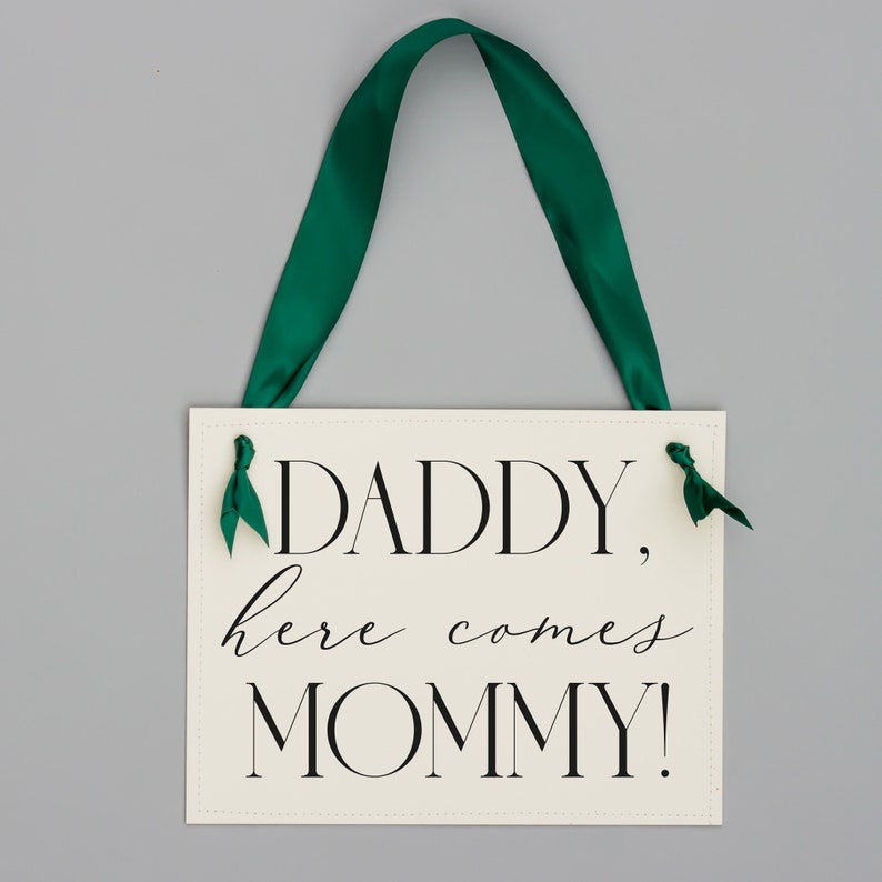May include: White sign with black text that reads "Daddy, here comes Mommy!" with green ribbon attached to the top.
