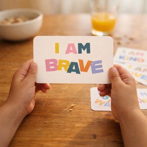 May include: A person holding a white card with the words "I AM BRAVE" in colorful block letters. Other cards with positive affirmations are visible in the background. A bowl of cereal and a glass of orange juice are also in the background.