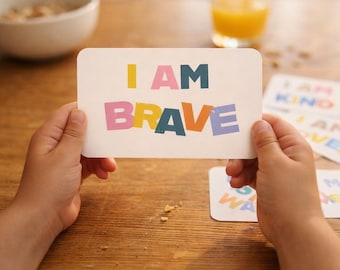 May include: A person holding a white card with the words "I AM BRAVE" in colorful block letters. Other cards with positive affirmations are visible in the background. A bowl of cereal and a glass of orange juice are also in the background.