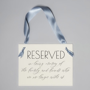May include: A cream-coloured sign with the word "RESERVED" in black, elegant font. Below, script reads "in loving memory of the family and friends who are no longer with us." A light blue ribbon is attached.