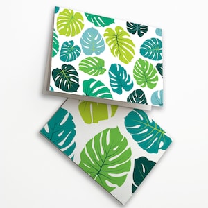 May include: Two greeting cards featuring a white backdrop and a repeating pattern of green and blue monstera leaves. The leaves are in various shades of green and blue, creating a tropical theme. One card is open, the other is closed.