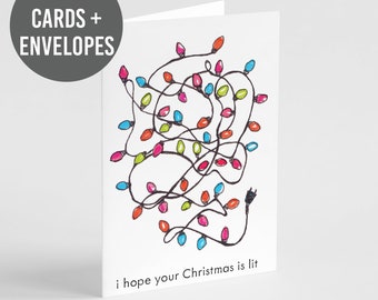 May include: A white greeting card featuring a hand-drawn illustration of tangled Christmas lights in red, blue, and green. The text at the bottom reads, i hope your Christmas is lit. A circular badge in the corner states 24 CARDS + ENVELOPES.