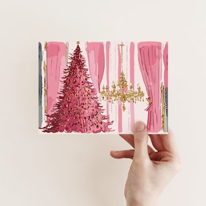 May include: A rectangular greeting card featuring a stylized illustration of a vibrant pink Christmas tree decorated with gold accents. The scene includes a gold chandelier and pink curtains against a light background, creating a festive interior setting.