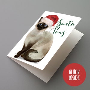 May include: A folded greeting card featuring a watercolor illustration of a Siamese cat wearing a Santa hat. The card has the text "Santa Paws" in green script. The inside of the card is blank.