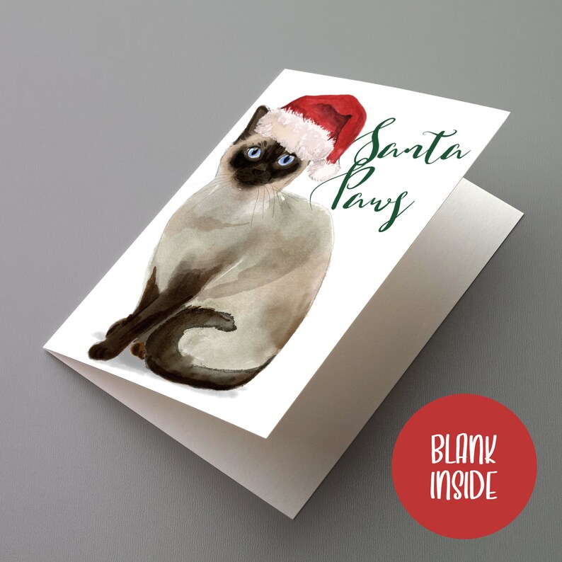 May include: A folded greeting card featuring a watercolor illustration of a Siamese cat wearing a Santa hat. The card has the text "Santa Paws" in green script. The inside of the card is blank.