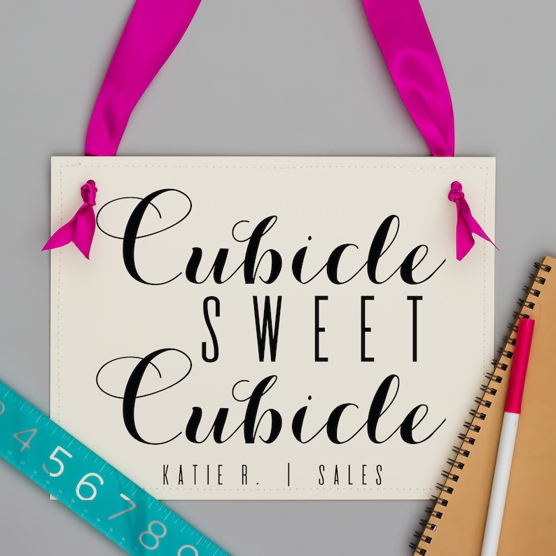 May include: White sign with black cursive text that reads "Cubicle Sweet Cubicle" and "Katie R. | Sales". The sign is hanging from pink ribbon.
