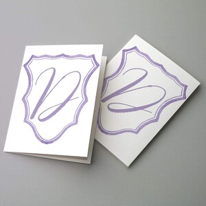 Può includere: Due biglietti pieghevoli bianchi con il contorno di uno stemma dipinto a mano color lavanda. Il biglietto a sinistra mostra una grande lettera D in corsivo, mentre il biglietto a destra mostra una grande lettera B in corsivo dello stesso tono.
