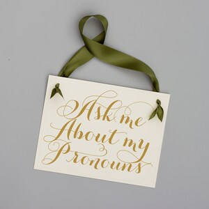 Puede incluir: Un cartel blanco con letras doradas que dice "Ask me about my pronouns" colgado de una cinta verde.