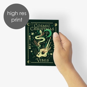 May include: A dark green print with the words "Cosmic Christmas Vibes" and illustrations of a snake, skull, candles, and celestial elements. The print is held by a hand against a white background.