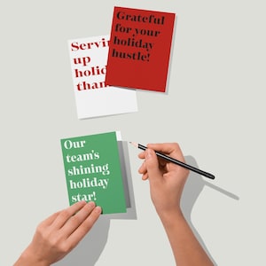 May include: A person writing on a green card with the text "Our team's shining holiday star!" with a pencil. Two other cards are visible, one red with "Grateful for your holiday hustle!" and one white with "Servir up holiday than."