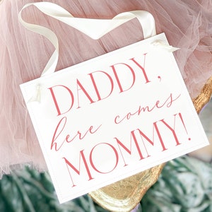 May include: White sign with pink text that reads "Daddy, here comes Mommy!"