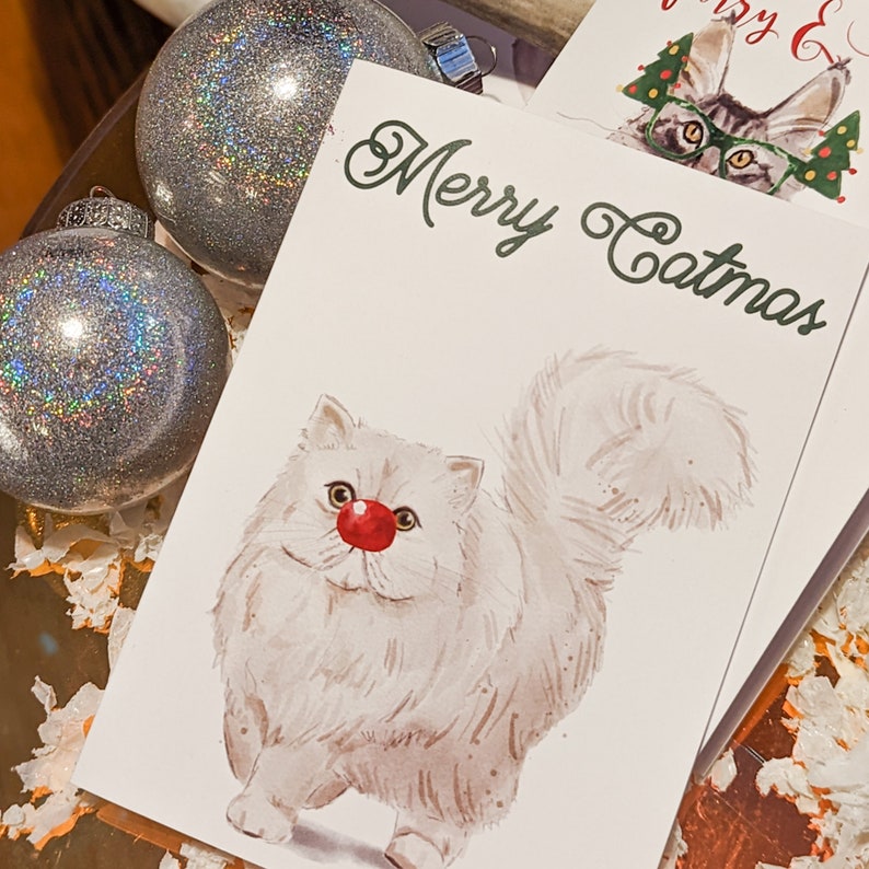 May include: Christmas cards with a watercolor illustration of a white cat with a red nose and the words "Merry Catmas" in green. Two silver glitter ornaments are in the background.