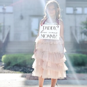 May include: A young girl in a pink tulle dress holds a sign that reads "Daddy, here comes Mommy!" The sign is white with black lettering and has a burgundy ribbon.