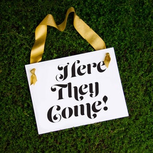 Puede incluir: Un cartel blanco con texto negro que dice "Here They Come!" colgado de una cinta dorada sobre un fondo de musgo verde.
