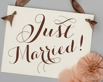 Op de afbeelding: Een wit bord met een bruin lint dat aan de bovenste hoeken is bevestigd. Het bord heeft de tekst "Just Married!" in bruine cursieve letters.