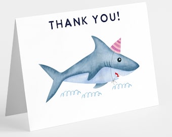 May include: A white folded greeting card featuring a watercolor illustration of a blue shark wearing a pink and white striped party hat. The shark has a red mouth and is swimming above blue curly lines. The text THANK YOU! is printed in blue.