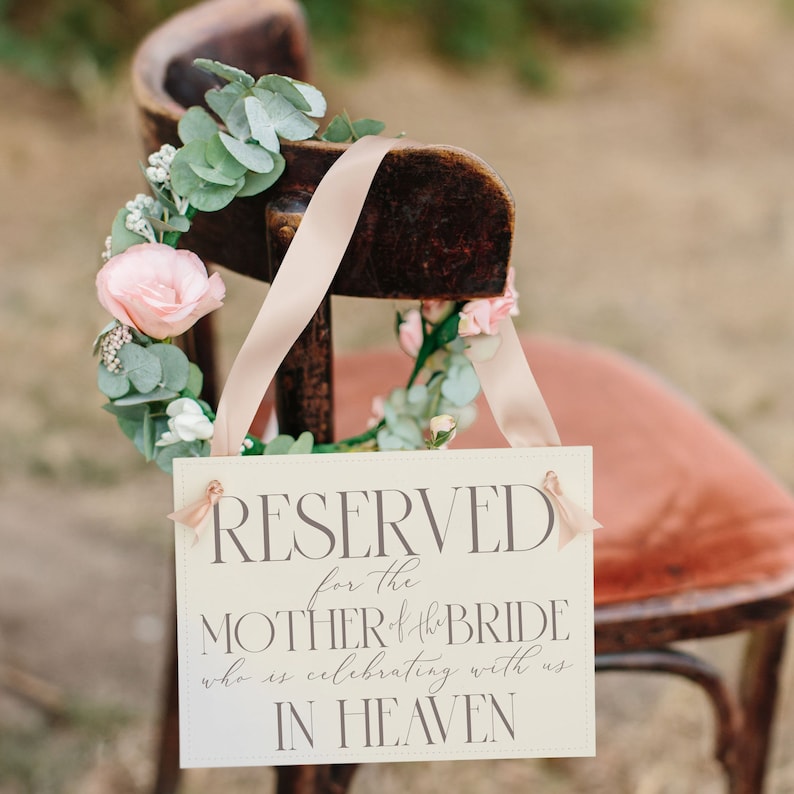 May include: A white sign with the text "RESERVED for the MOTHER of the BRIDE who is celebrating with us IN HEAVEN" hanging on a wooden chair decorated with a floral wreath.