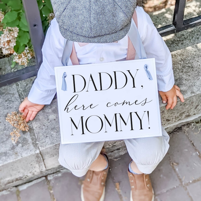 May include: A white sign with black text that reads "Daddy, here comes Mommy!"  A young child in a white shirt, grey pants, and a grey cap holds the sign.