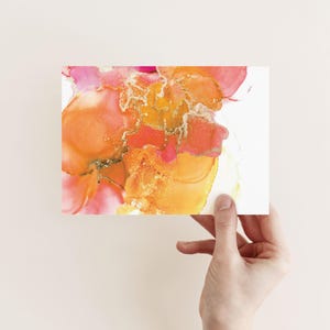 A hand is holding up a piece of paper with an abstract, colorful painting of flowers or flowers in a liquid medium.