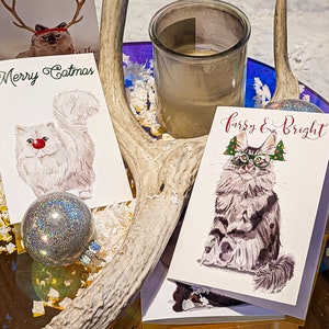 May include: Christmas-themed greeting cards featuring watercolor cat illustrations. One card reads "Merry Catmas" with a white cat wearing a red nose. Another card says "Furry & Bright" with a cat wearing glasses and Christmas tree decorations. A silver glitter ornament is also present.