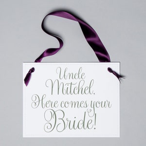 May include: A white rectangular sign with the text "Uncle Mitchel, Here comes your Bride!" in elegant script. The sign is adorned with a deep purple ribbon for hanging, adding a touch of sophistication to the wedding accessory.