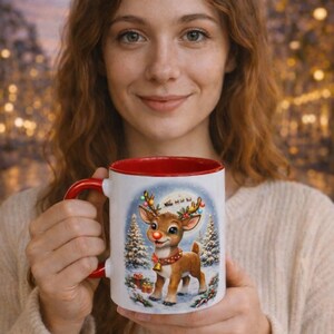Op de afbeelding: Witte keramische mok met rode binnenkant en oor, met een feestelijke illustratie van een rendier met een rode neus, met een belletjesketting, in een besneeuwde scène. Kerstthema.