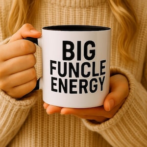 Puede incluir: Taza de cerámica blanca con interior y asa negros, con el texto "BIG FUNCLE ENERGY" en letras negras en negrita. La taza la sostiene una persona que lleva un jersey beige.