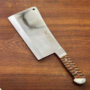 May include: A stainless steel cleaver with a polished blade and a patterned orange and brown handle. The cleaver has the text "LUCKY" on the blade. The cleaver is set against a wooden surface.