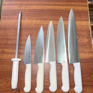 May include: A set of six stainless steel kitchen knives with white handles, ranging in size, and a sharpening steel. The knives are arranged on a wooden surface, showcasing their blades and handles.